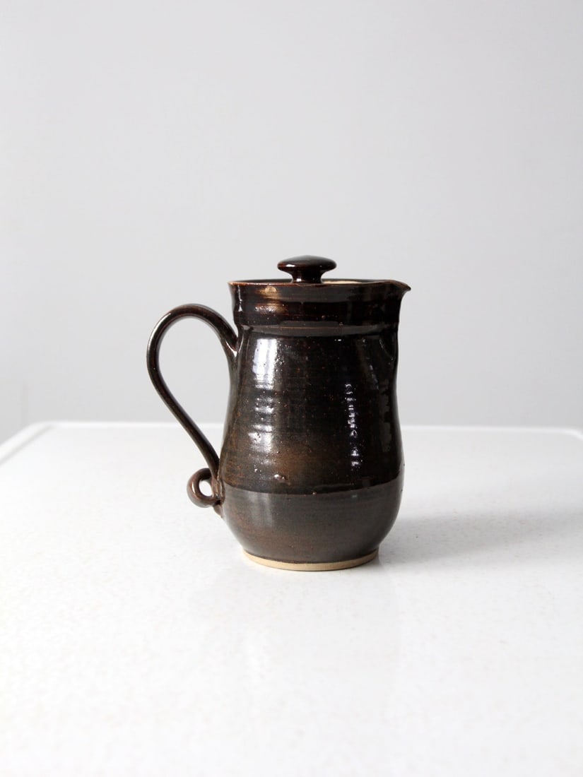 1978 Vintage Studio Pottery Pitcher Dark Brown Signed: 1978 Vintage Studio Pottery Pitcher Dark Brown Signed This vintage studio pottery pitcher, crafted in 1978, showcases a rich dark brown ceramic finish with a uniquely curled handle and an inset lid. T