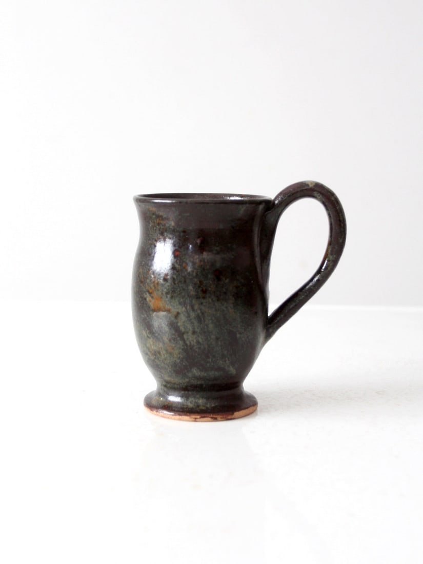 Artisan Vintage Studio Pottery Mug Brown Green Glaze 5.25": Artisan Vintage Studio Pottery Mug Brown Green Glaze 5.25" This vintage handmade studio pottery mug showcases a softly rounded body with a curved handle and a flared foot. The unique glaze features a