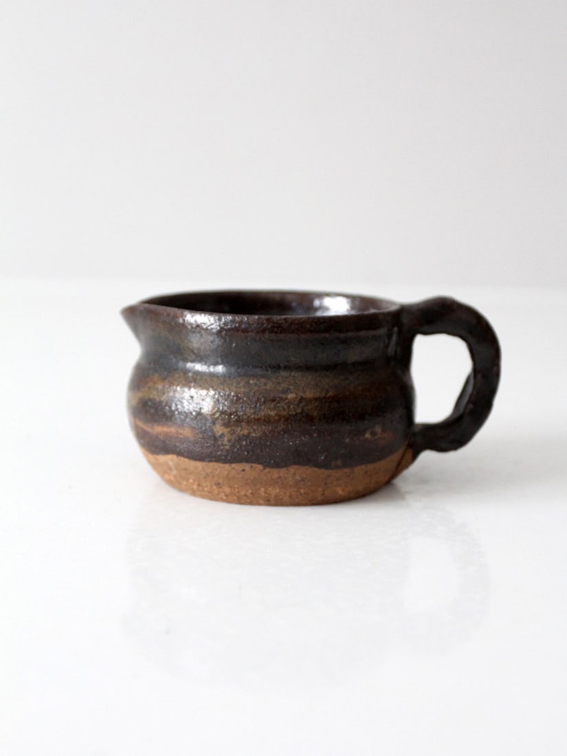 Artisanal Vintage Studio Pottery Creamer Deep Brown Glaze: Artisanal Vintage Studio Pottery Creamer Deep Brown Glaze This vintage studio pottery creamer showcases a unique hand-shaped form that emphasizes its artisanal quality. The deep brown dipped glaze bea