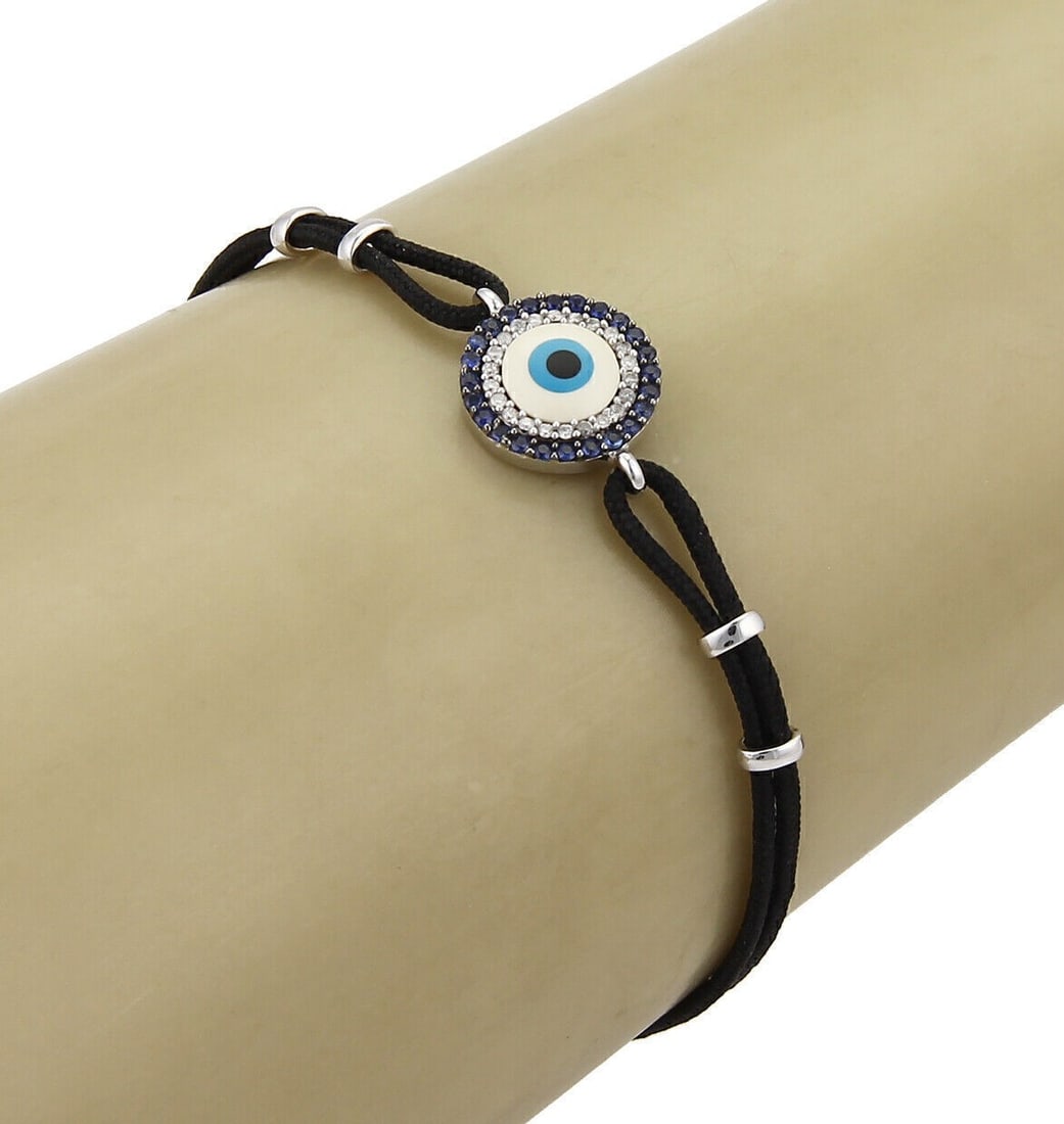 Cord Bracelet with 18k White Gold Evil Eye Sapphire and Diamond Charm: Cord Bracelet with 18k White Gold Evil Eye Sapphire and Diamond Charm This 18k white gold cord bracelet features an evil eye charm adorned with 0.10ct of diamonds and 0.20ct of sapphires. The bracelet