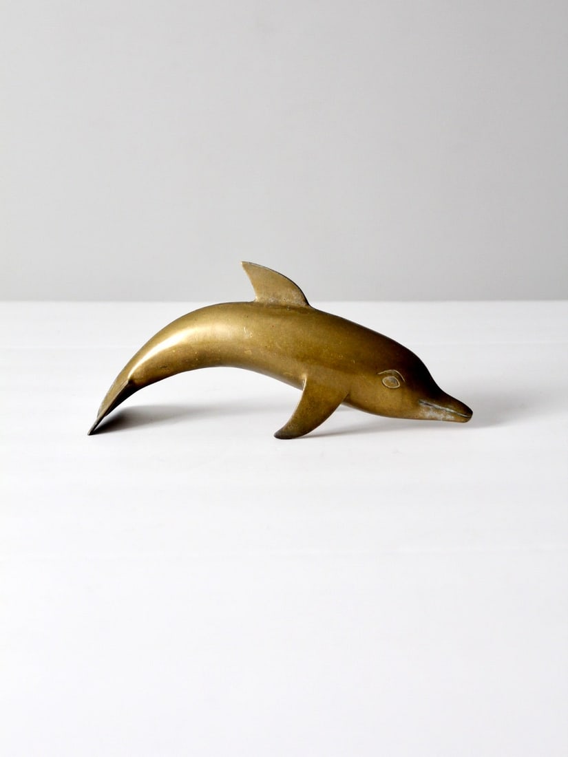 Vintage Mid-Century Brass Dolphin Figurine Coastal Decor: Vintage Mid-Century Brass Dolphin Figurine Coastal Decor This vintage mid-century brass dolphin figurine showcases a smooth curved body, arched fins, and expressive facial details. It serves as a char