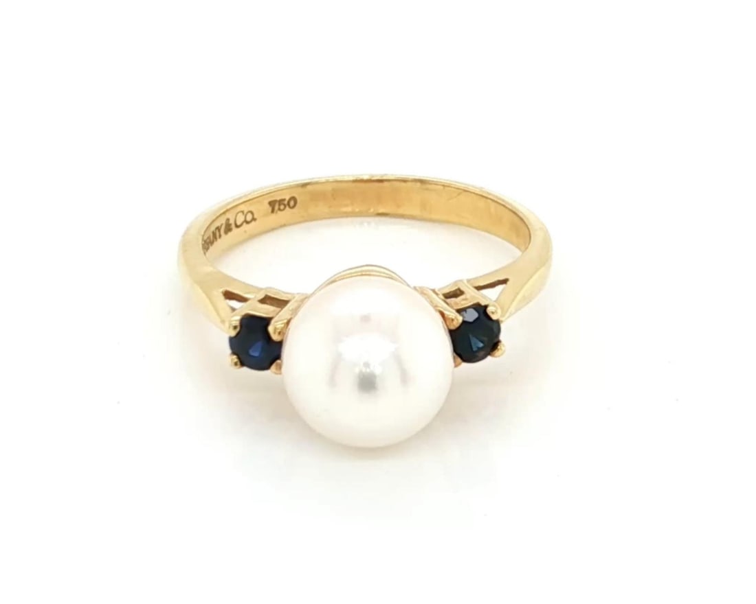 18k Gold Tiffany Pearl and Sapphire Ring Size 5: 18k Gold Tiffany Pearl and Sapphire Ring Size 5 This Tiffany & Co. ring is crafted from solid 18k yellow gold and features a central 8 mm pearl complemented by sapphire accents. The ring is designed i