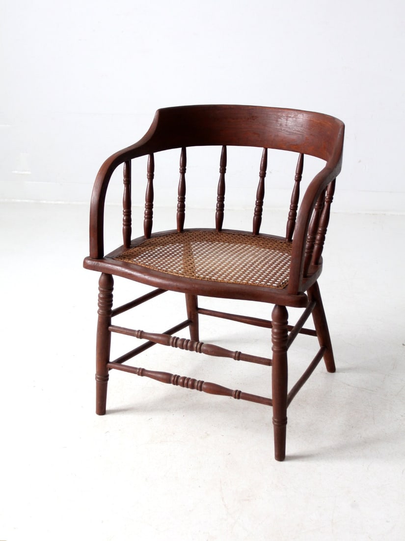 Classic Antique Captain's Chair with Cane Seat and Spindle Details: Classic Antique Captain's Chair with Cane Seat and Spindle Details This antique captain's chair showcases a classic barrel-back silhouette complemented by intricate turned spindle detailing. The chair