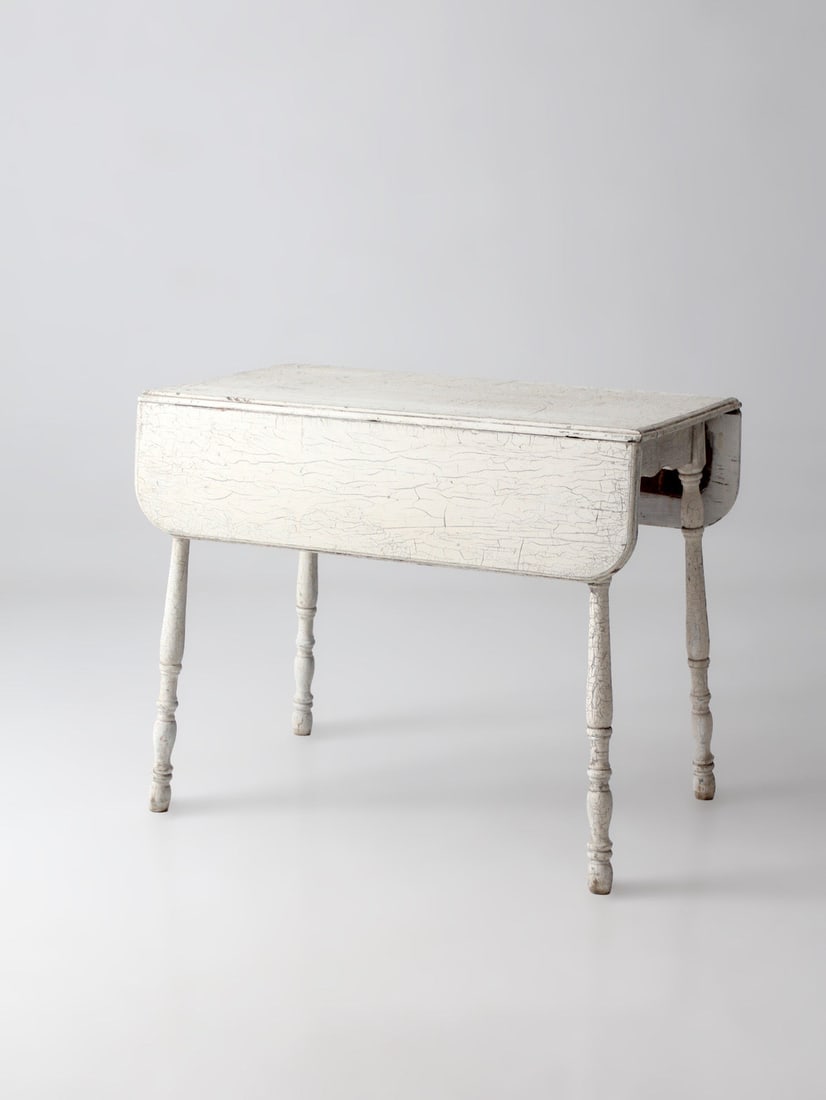 White Painted Antique Drop Leaf Table with Turned Legs: White Painted Antique Drop Leaf Table with Turned Legs Antique white painted drop leaf farmhouse table with a slim rectangular form, turned supports, and softly curved aprons. It features a weathered 