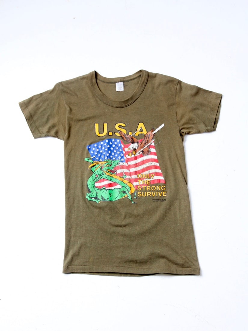 1987 Vintage USA Graphic Tee Eagle Dragon Design: 1987 Vintage USA Graphic Tee Eagle Dragon Design Discover a piece of the 1980s with this Vintage USA Graphic Tee. This ultra-soft olive green t-shirt features a striking design of an eagle and a drago