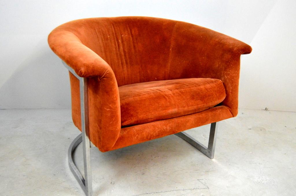 Milo Baughman Style Club Chair: With a shaped back and arms upholstered in burnt orange resting in a chrome body. 27 x 33 x 30 in. Note: Fabric is worn and the point of attachment on the rear of the chair is loose.