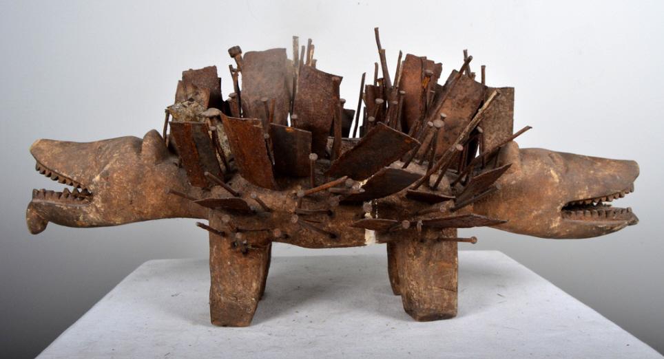 Bakongo Dog Fetish Figure: African, Congo, Nkisi Nkonde. wood and metal; Of a double headed dog, with a bilongo at the center and decorated with metal shards and nails, each head with mouth open and tongue hanging out. 12 x 26