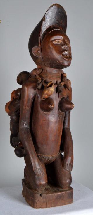 Modern West African Figure of a Female Shaman: West African, 20th Century, wood; of a kneeling woman with a necklace comprised of whelk shells, gourds and a turtle shell. 38 x 8 x 9 in. Condition: Wear consistent with age and use Property of Antho