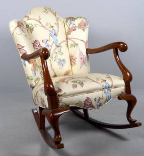 Queen Anne Style Rocking Armchair: With an upholstered and paneled back, the upholstered seat flanked by shaped and carved arms with scrolling stumps, the whole raised on two cabriole legs ending in pad feet to the front and two splay