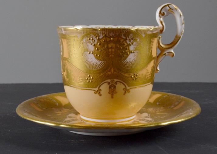 Royal Worcester Demitasse Cup and Saucer: The encrusted gold landscape is on beige ground with the inside of the cup a white porcelain . The saucer is encrusted with the gold leaf landscape and the back is white. Cup 3 in. D x 3in. Saucer 4.5