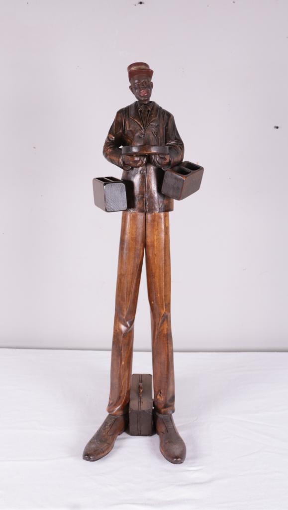 Folk Art Carved Delivery Man: Carved statue of an African American delivery man carrying a tray and two wooden boxes, with a suitcase between his legs. Crank on back turns but nothing happens. Dimensions: 36 x 12 x 10 in. NOTE: