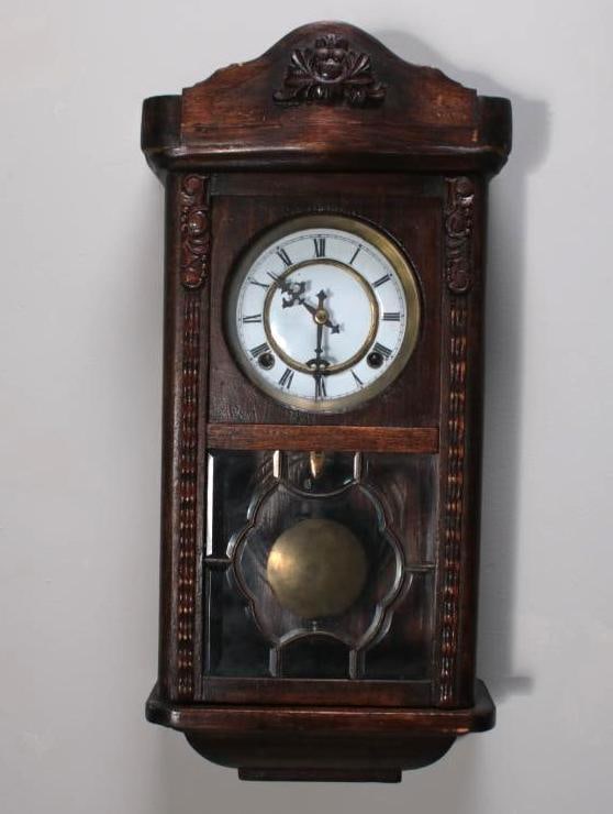 Walnut Wall Clock: With a carved and glazed case over an enammeled, roman face. Keys and works present. Marked "LOC | CK" inside. Dimensions: H 23 x W 10.5 x D 5 inches NOTE: Wear consistent with age and use