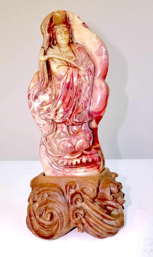 Carved Hardstone Figure of Guanyin on Stand: Carved hardstone figure depicting Guanyin standing in flowing robes, holding a ruyi-style scepter, the stone of variegated pink, red, and cream coloration with natural inclusions utilized in the carvi