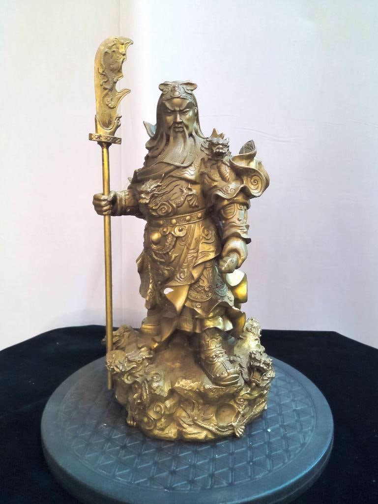 Chinese Figural Sculpture of Guan Yu (Kwan Kung): Cast resin figural sculpture depicting Guan Yu in armor holding a guan dao, standing on a rocky base with dragon motifs. Finished in a gilt-toned surface. Dimensions: H 10.5 x W 5 x D 5 inches NOTE: G