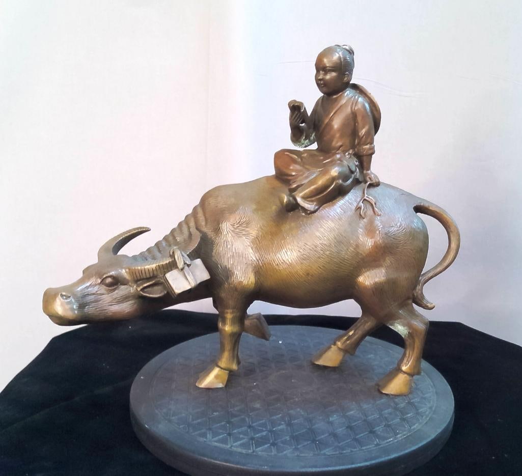 Chinese Figural Group of Boy on Water Buffalo: Cast resin figural group depicting a seated boy holding an object while riding a water buffalo, rendered with textured hide and a gilt-toned finish. Dimensions: H 9 x W 12 x D 9 inches NOTE: Surface w