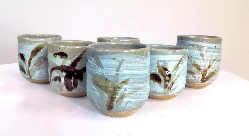 Set of Six Studio Pottery Stoneware Tea Cups: six hand-thrown stoneware tea cups (yunomi form), decorated with pale celadon-blue glazes and freehand botanical motifs. The cylindrical vessels display subtle variations consistent with handmade prod