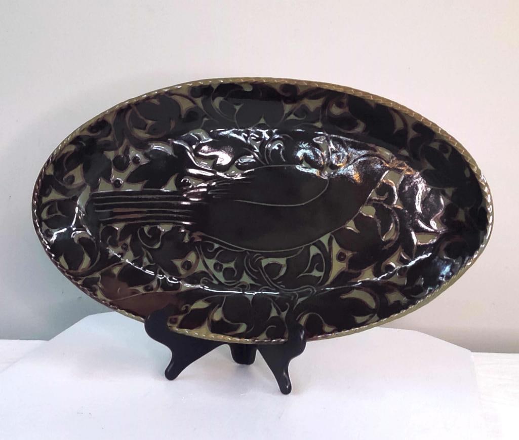Studio Art Pottery Bird Motif Oval Platter: Handcrafted studio pottery oval platter featuring a stylized bird in relief against a scrolling foliate ground. Finished in a glossy dark glaze with contrasting carved decoration. Raised footed base. 