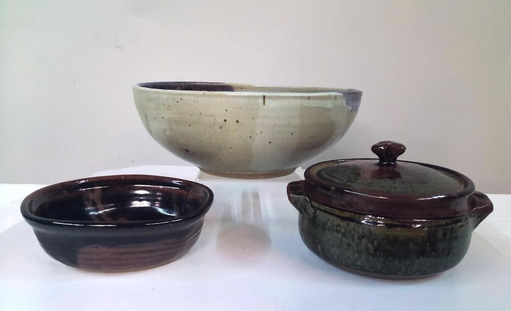 Three Studio Pottery Serving Vessels: Group of three hand-thrown studio pottery vessels comprising a large serving bowl with cream and purple glazed decoration, a covered casserole form with mottled green and brown glaze, and a shallow br