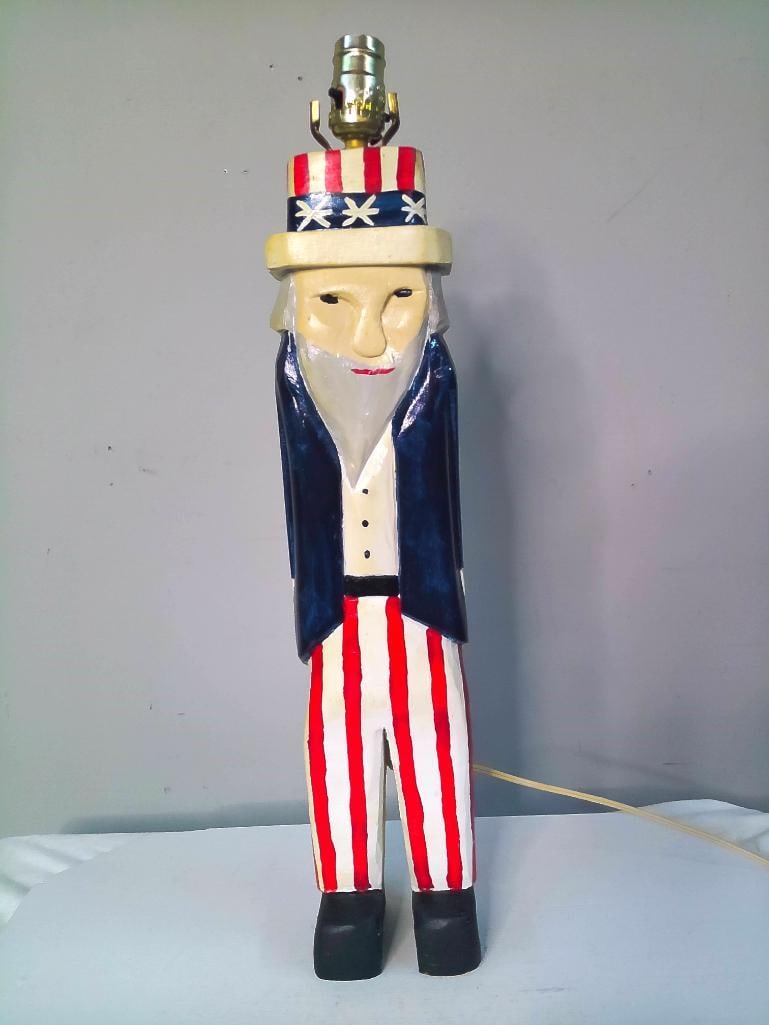 Initialed D.S.E., Uncle Sam Folk Art Table Lamp: 1998, Carved and polychrome-painted wooden figural lamp depicting Uncle Sam in a patriotic top hat, dark coat, white shirt, red-and-white striped trousers, and black shoes. Brass socket mounted atop t