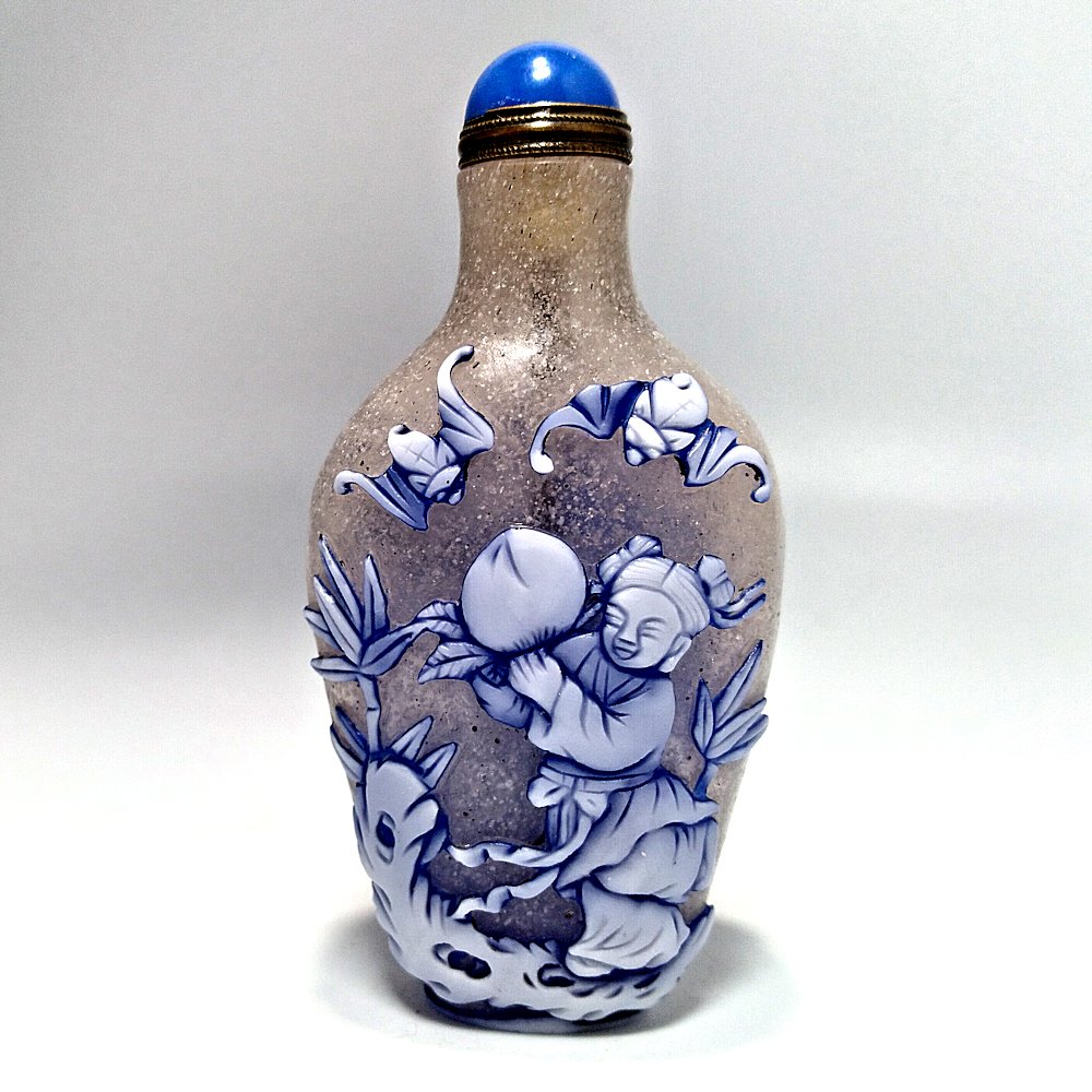 PEKING GLASS OVERLAY SNUFF BOTTLE FEMALE IMMORTAL: THE WELL CARVED BOTTLE DEPICTING A FEMALE IMMORTAL ON ONE SIDE AND A CHILD WITH A PEACH AND BATS, CARVED IN BLUE AND WHITE LAYERS OVER BUBBLE INFUSED SNOWFLAKE GLASS.