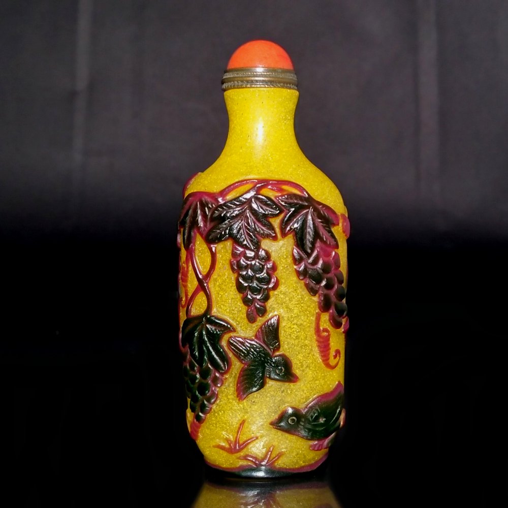 EXQUISITE YELLOW PEKING GLASS SNUFF BOTTLE 18-19TH C: THE BOTTLE OF YELLOW GLASS IS SO BEAUTIFULLY CARVED IN DEEP BROWN WITH SMOOTH LINES AND MUCH DETAIL. IT FEATURES DUCKS AND LOTUS AND CHICKS AND GRAPES. IT IS 3.5 INCHES TALL. PROVENANCE: MAJOR PRIVATE