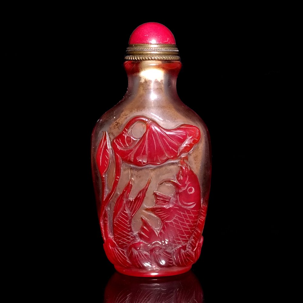 CHINESE RED GLASS SNUFF BOTTLE KOI FISH QING: The lovely bottle is carved in red over clear glass with much detail. It depicts Koi fish and lotus. The foot is surrounded by a wave pattern. The bottle is 3 inches tall.