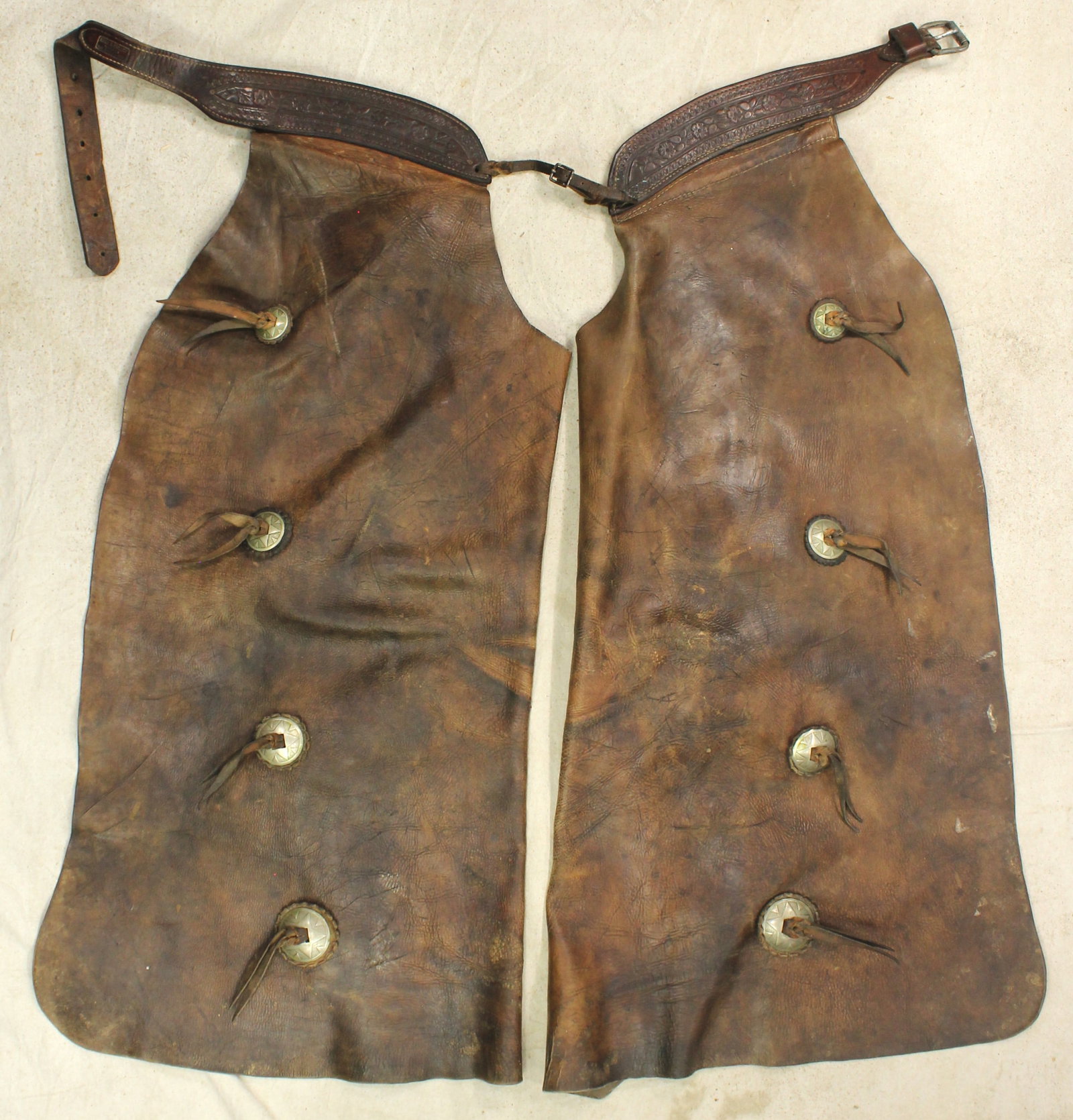 Kuck & Bonney Batwing Chaps: Heavy leather cowboy batwing chaps featuring saddle string conchos and uniquely tooled maker marked belt, from The Dalles Oregon. Exhibits substantial period use with rich patina throughout.