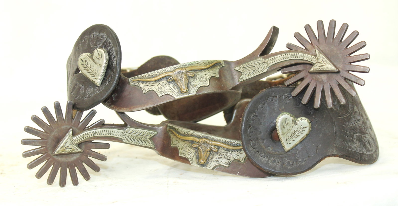 Bill Adamson Bull Head Spurs: Wonderful pair of double mounted Adamson spurs featuring a steerhead pattern, with card suits on the offside, oversized heart buttons, finished with arrow shanks and huge rowels. Completed by tooled