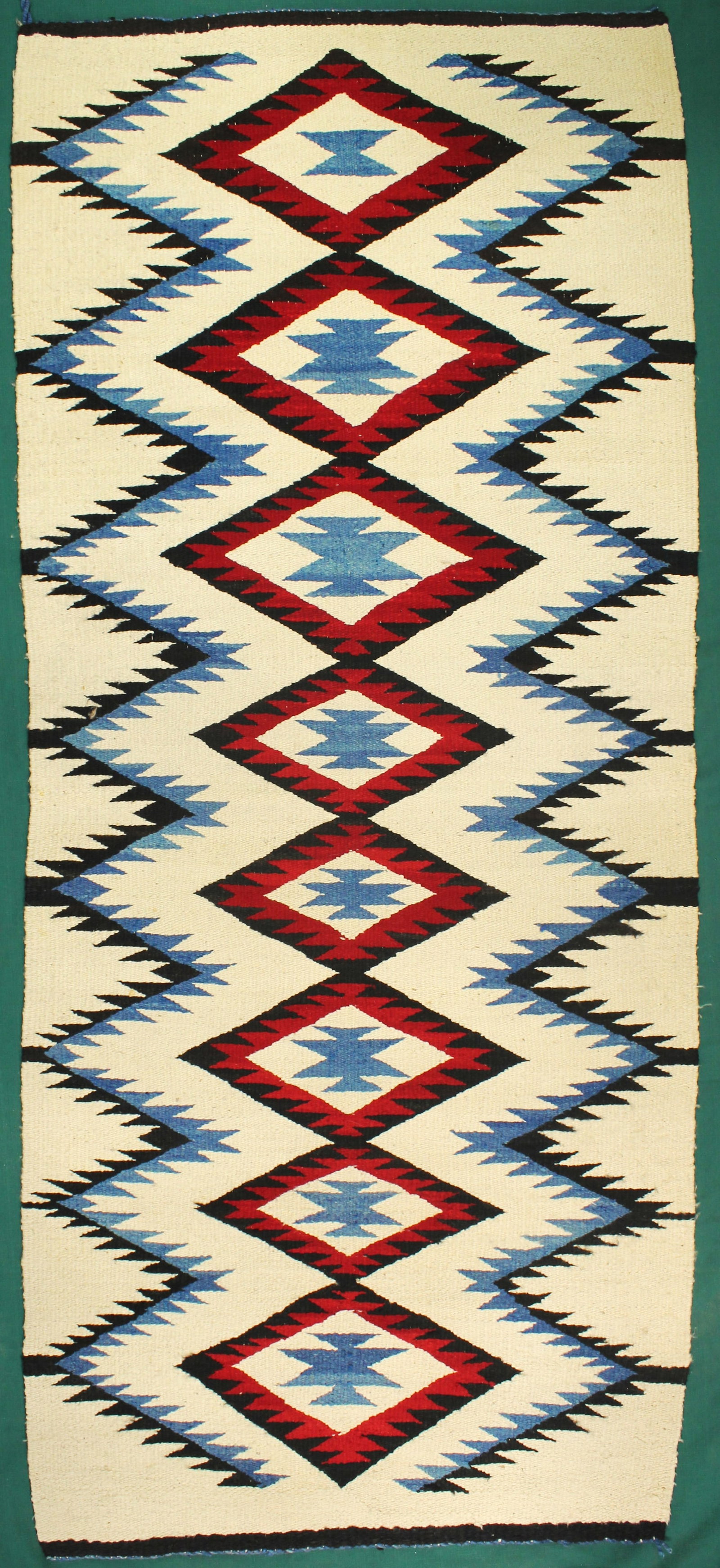 Navajo Red Mesa Runner: Handwoven Navajo runner featuring repeating diamond and serrated geometric motifs in red, black, white, and blue. Fine decorative weaving suitable for wall or floor display. Dimensions 31”x