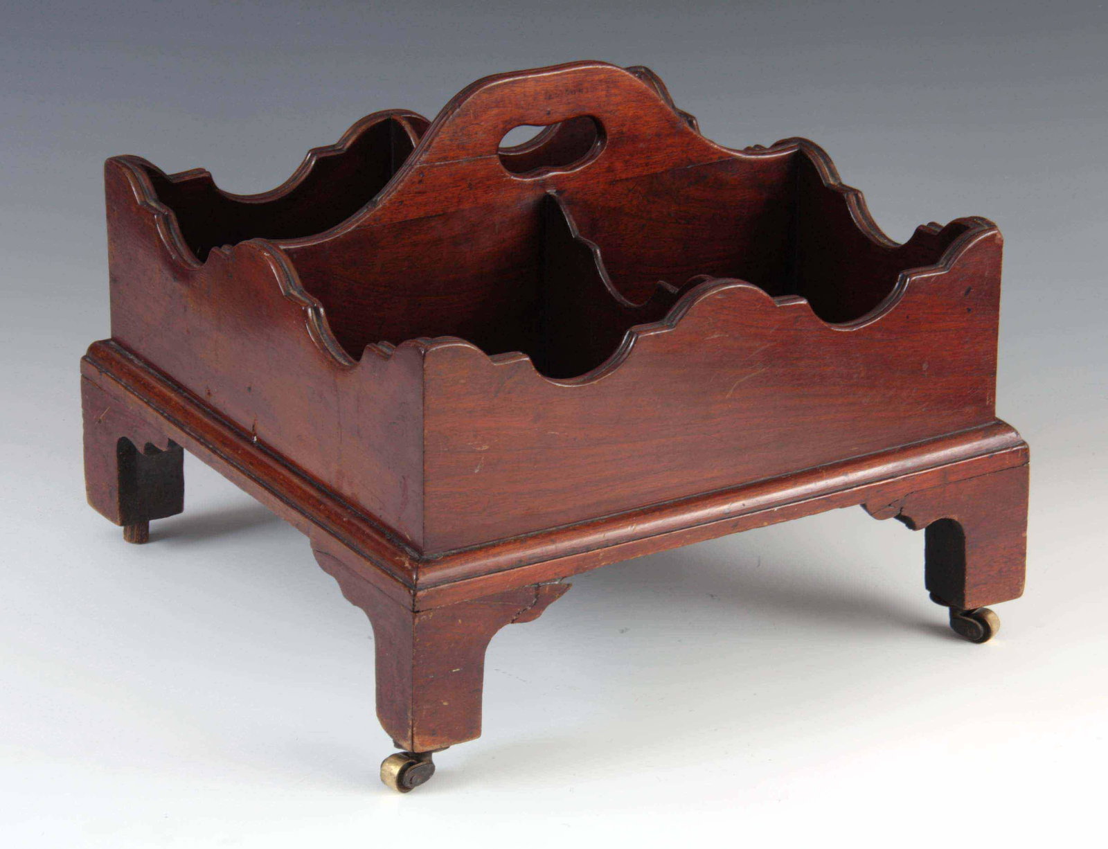 A GEORGE III FIGURED MAHOGANY TABLE TOP BUTLERS DE: A GEORGE III FIGURED MAHOGANY TABLE TOP BUTLERS DECANTER/BOTTLE CARRIER of square form with shaped sides joined by a carrying handle; standing on small bracket feet with brass casters 35cm square 28cm
