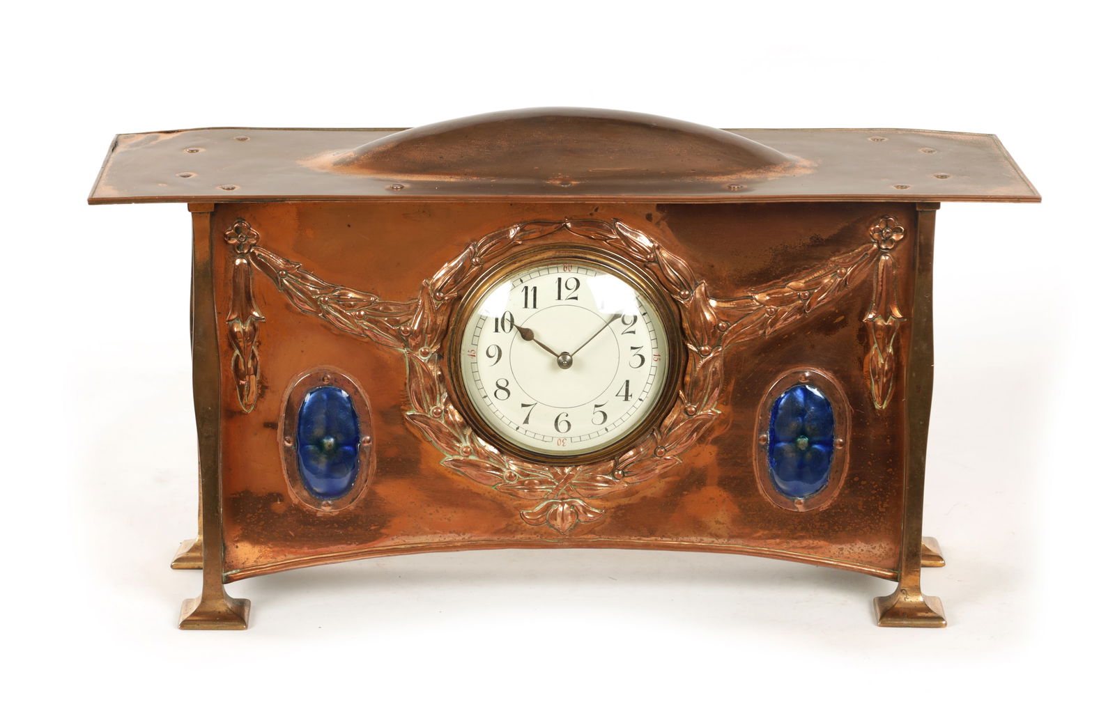 ATT. GEORGE WALTON. A COPPER AND ENAMEL ARTS AND CRAFTS MANTEL CLOCK: ATT. GEORGE WALTON. A COPPER AND ENAMEL ARTS AND CRAFTS MANTEL CLOCK the riveted top with domed centre above an enamel Arabic dial surrounded by a relief laurel leaf garland and two blue enamel caboch