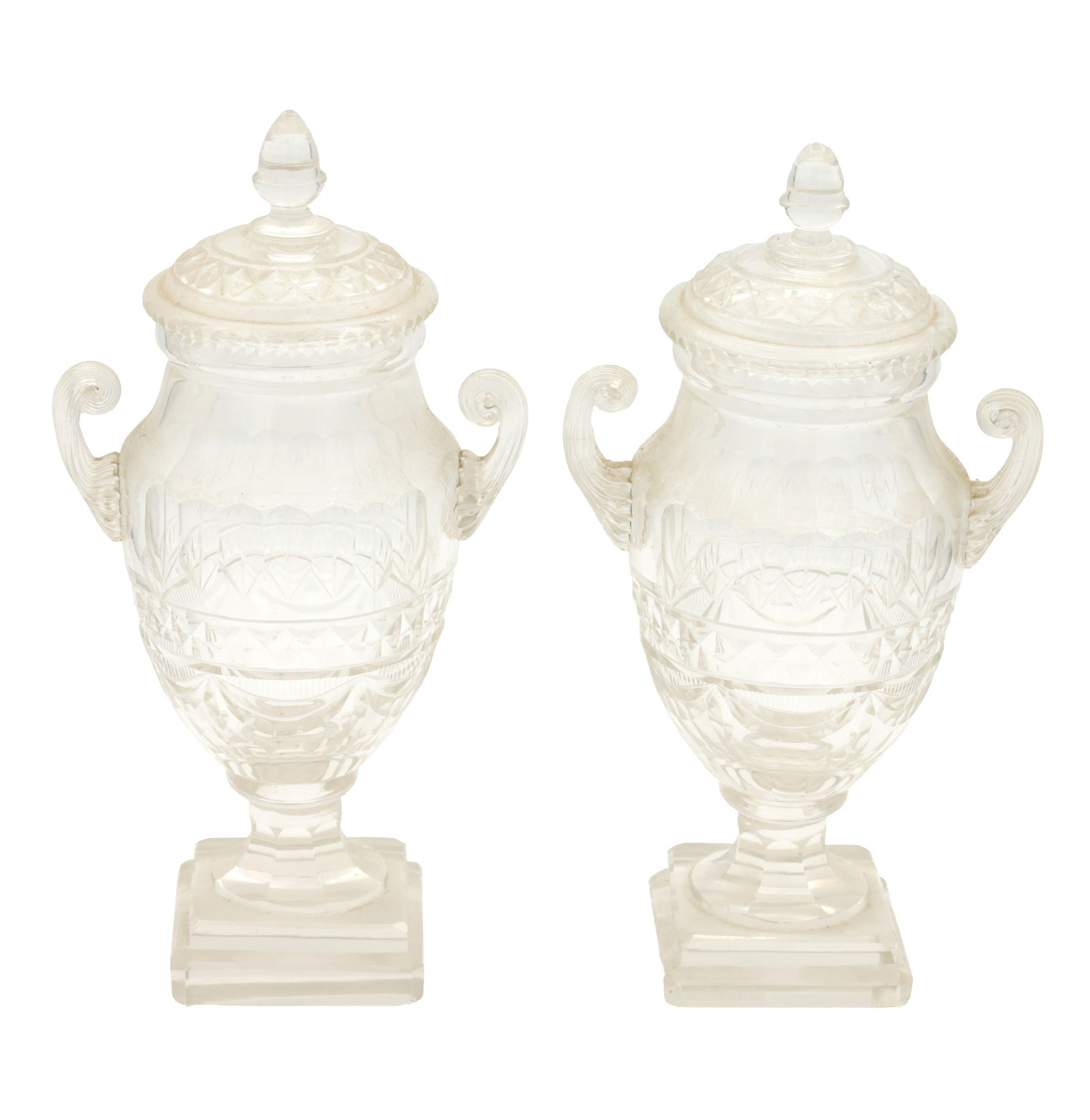 A PAIR OF REGENCY CUT GLASS LIDDED URNS: A PAIR OF REGENCY CUT GLASS LIDDED URNS with scolled side handles and raised on square platform bases. (26cm high )