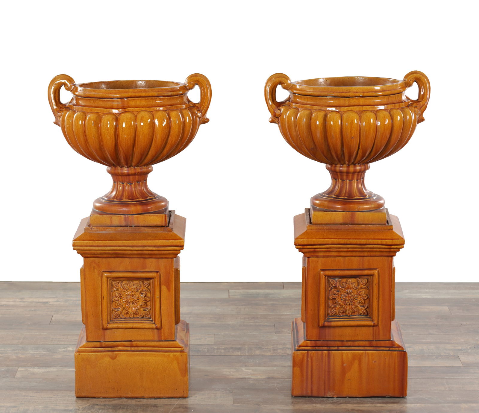 A PAIR OF 19TH CENTURY MAJOLICA GLAZED POTTERY GARDEN URNS ON PEDESTALS: A PAIR OF 19TH CENTURY MAJOLICA GLAZED POTTERY GARDEN URNS ON PEDESTALS each of campana form with twin loop handles and lobed bodies in a mottled brown glaze, raised on waisted circular stems and squa