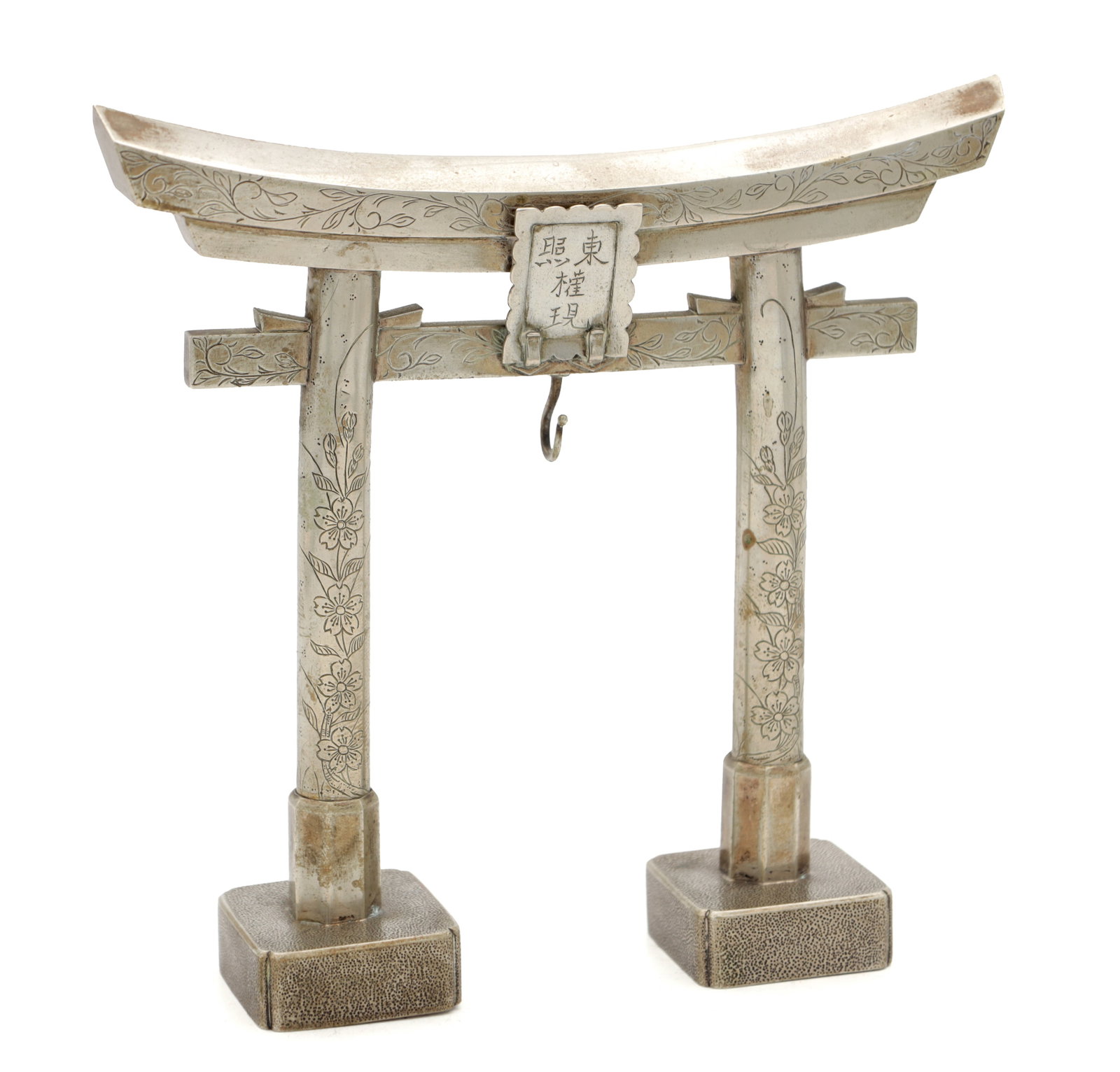A MEIJI PERIOD JAPANESE SILVER METAL POCKET WATCH HOLDER FORMED AS A TORII SHINTO GATE: A MEIJI PERIOD JAPANESE SILVER METAL POCKET WATCH HOLDER FORMED AS A TORII SHINTO GATE with engraved floral decoration, a central suspension hook beneath an inscribed plaque, on twin textured square b
