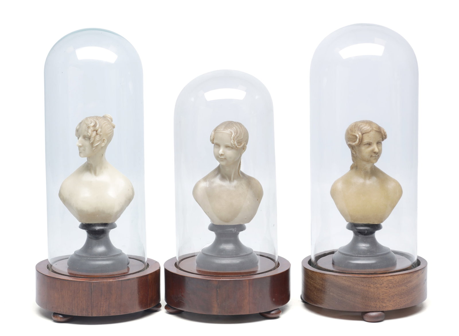 A COLLECTION OF THREE WAXWORK BUSTS OF YOUNG LADIES UNDER GLASS DOMES ON MAHOGANY BASES: A COLLECTION OF THREE WAXWORK BUSTS OF YOUNG LADIES UNDER GLASS DOMES ON MAHOGANY BASES each modelled with classical coiffures and mounted on ebonised turned socles beneath tall cylindrical domes set
