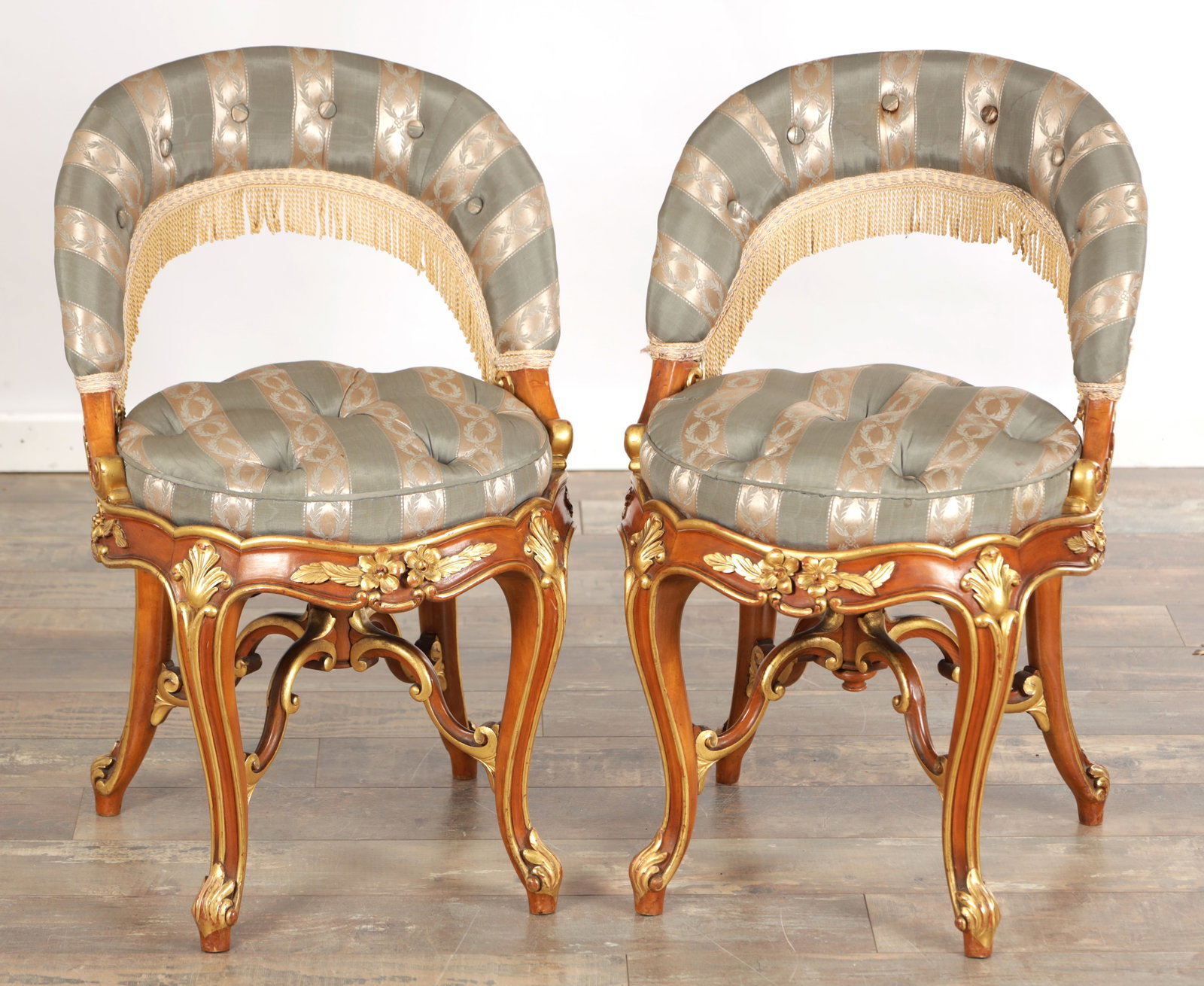 A PAIR OF 19TH CENTURY STYLE FRENCH WALNUT AND PARCEL GILT UPHOLSTERED DRESSING TABLE CHAIRS: A PAIR OF 19TH CENTURY STYLE FRENCH WALNUT AND PARCEL GILT UPHOLSTERED DRESSING TABLE CHAIRS of tub back form with tasselled fringe and deep buttoned striped seats, the shaped aprons carved with flora