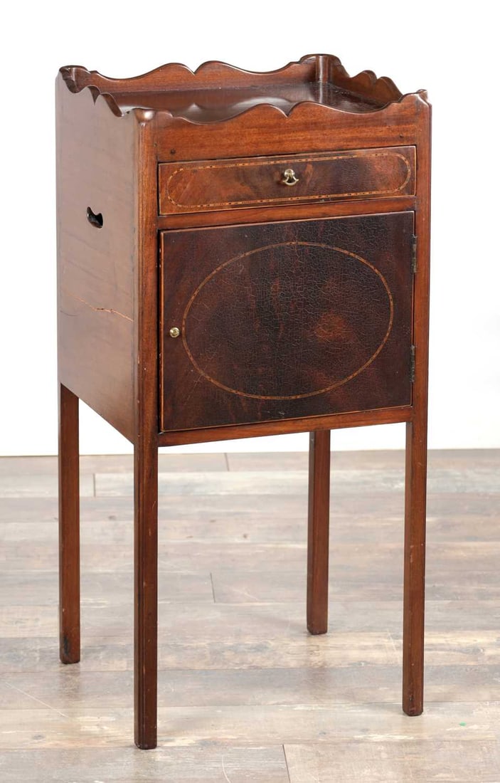 A GEORGE III MAHOGANY BEDSIDE COMMODE: A GEORGE III MAHOGANY BEDSIDE COMMODE with a three-quarter shaped gallery above a line-inlaid short drawer and an oval-inlaid cupboard door with brass knob pulls and side carrying handles, raised on