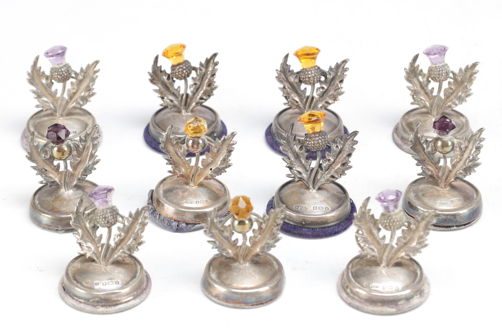 A SET OF ELEVEN SCOTTISH SILVER THISTLE MENU HOLDERS: A SET OF ELEVEN SCOTTISH SILVER THISTLE MENU HOLDERS each formed as an openwork thistle spray set with faceted coloured stones, on circular stepped bases with hallmarks to the bases, several with
