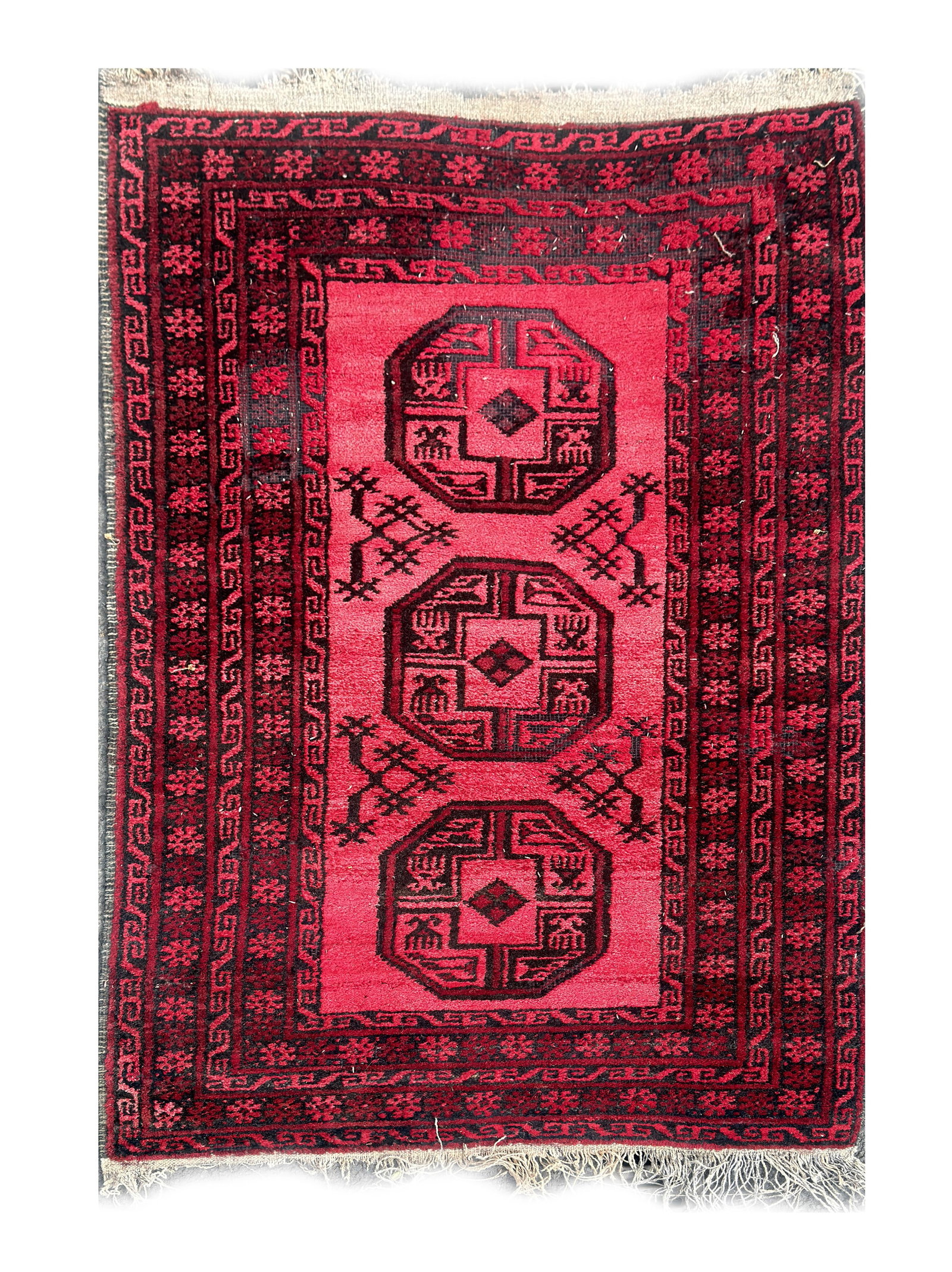 AN AFGHAN AQCHA WOOL CARPET: AN AFGHAN AQCHA WOOL CARPET of rectangular form with a madder red field centred by three large octagonal guls and hooked devices within multiple geometric borders, fringed ends. (185cm by 107cm )