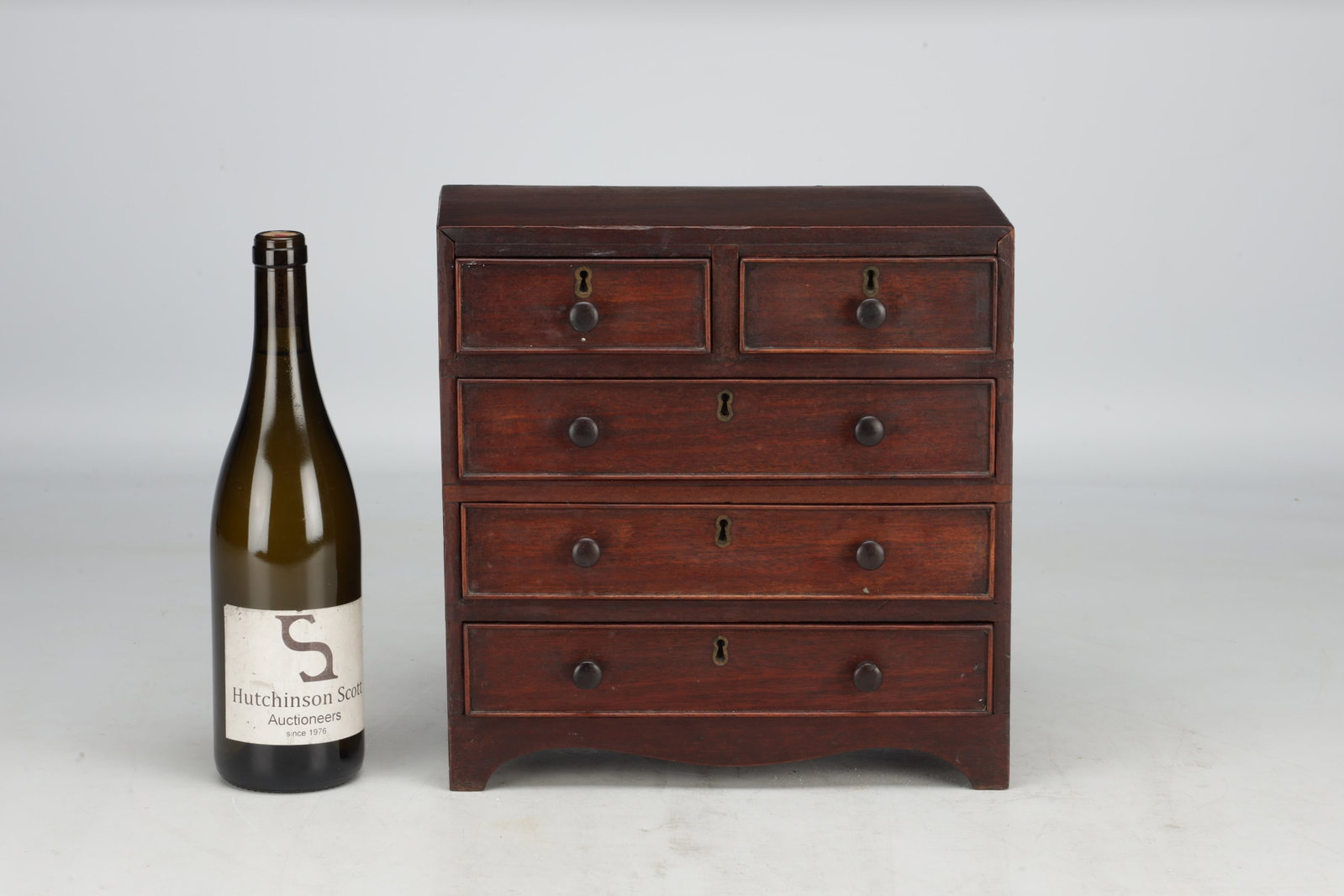 A 19TH CENTURY MAHOGANY MINIATURE CHEST OF DRAWERS: A 19TH CENTURY MAHOGANY MINIATURE CHEST OF DRAWERS with rectangular top above two short over three long drawers fitted with turned wooden knobs and brass escutcheons, on a shaped apron and bracket