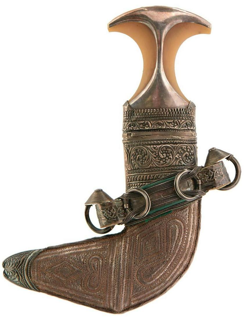 AN EARLY 20TH CENTURY OMANI JAMBIYA, 18cm bright: AN EARLY 20TH CENTURY OMANI JAMBIYA, 18cm bright sharply curved blade with raised medial ridge, characteristic white metal mounted hilt decorated with scrolling foliage, contained in its white metal m
