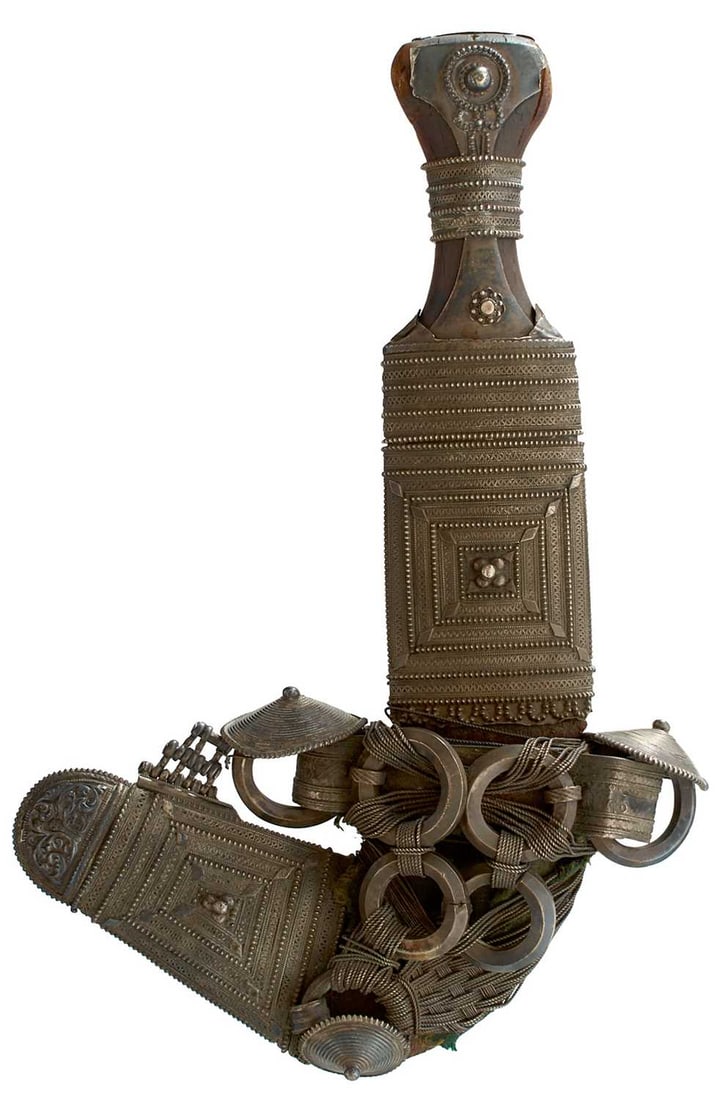 A 19TH CENTURY OMANI JAMBIYA,: 19cm sharply curved blade with raised medial ridge, characteristic white metal mounted hilt with filigree decoration and horn grip, in its leather wrapped wooden sheath with ornate white metal