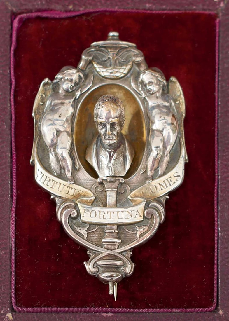 A DUKE OF WELLINGTON MEMORIAL BROOCH,: central bust of Wellington flanked by cherubs and surmounting the motto VIRTUTIS FORTUNA COMES in scroll banner form, pin backed, in its burgundy velvet and silk lined case. The back marked PUBD. BY