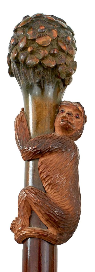 AN EARLY 20TH CENTURY WALKING STICK,: the carved wood handle in the form of a monkey climbing a flowering stump, white metal collar, tapering wooden haft, horn ferrule, 92cm long over all.
