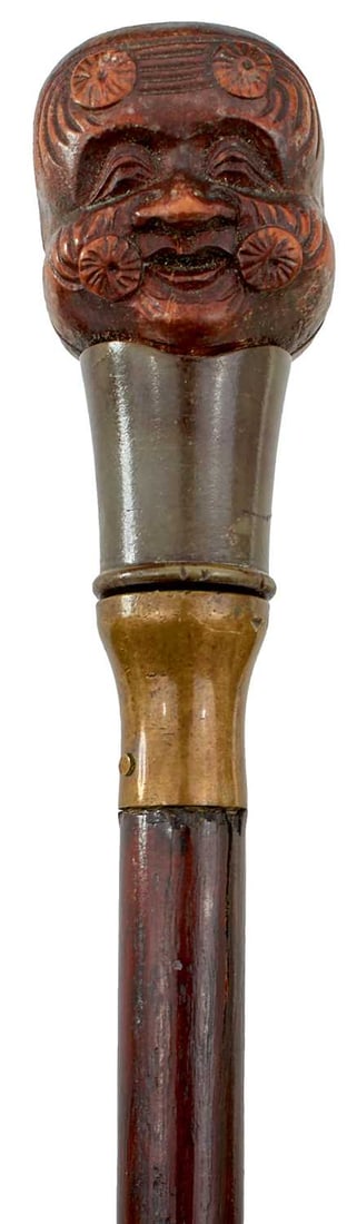 A JAPANESE WALKING STICK,: the carved wood handle of netsuke form decorated with the face of a baby opposing the face of an old man with mon, metal collar, slender tapering haft (split), no ferrule, 98.5cm long over all.
