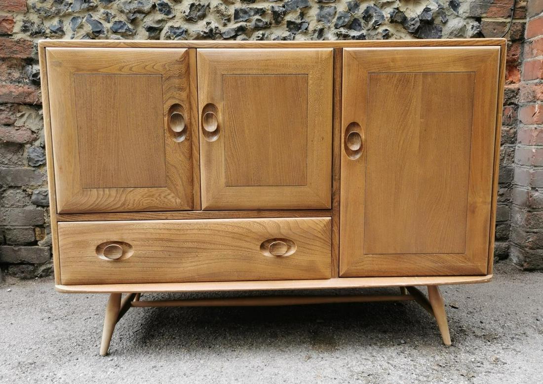 A small blonde elm Ercol sideboard, with twin cupboard: A small blonde elm Ercol sideboard, with twin cupboard doors above a single frieze drawer, flanked with a shelved cupboard, supported on four turned legs with stretcher, paper label to inside of right
