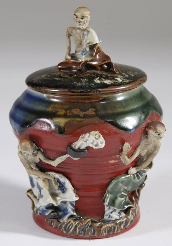 Early 20th C. Sumida Gawa Covered Tobacco Jar: Applied half naked figures of two men with one holding a incense censor and the other waving his hand at it. Applied half naked figure to lid of man sitting. 8" H; applied signed cartouche on back of