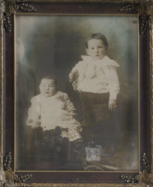 Large Antique Photo of Children: Depicting a baby sitting in a chair and his/her brother standing on a chair beside him/her. Both in period clothing. Photo has had color added. Framed behind glass, and bottom right corner is missing.