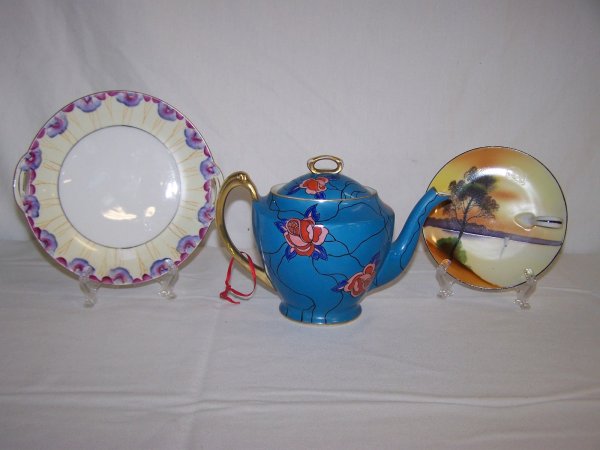 Lot Of Three Noritake Hand Painted Table Articles: Lot includes blue hand painted teapot with red and pink flowers outlined in black with gold hand painted handle and trim along with a hand painted finger hole plate and a two handled floral decorated