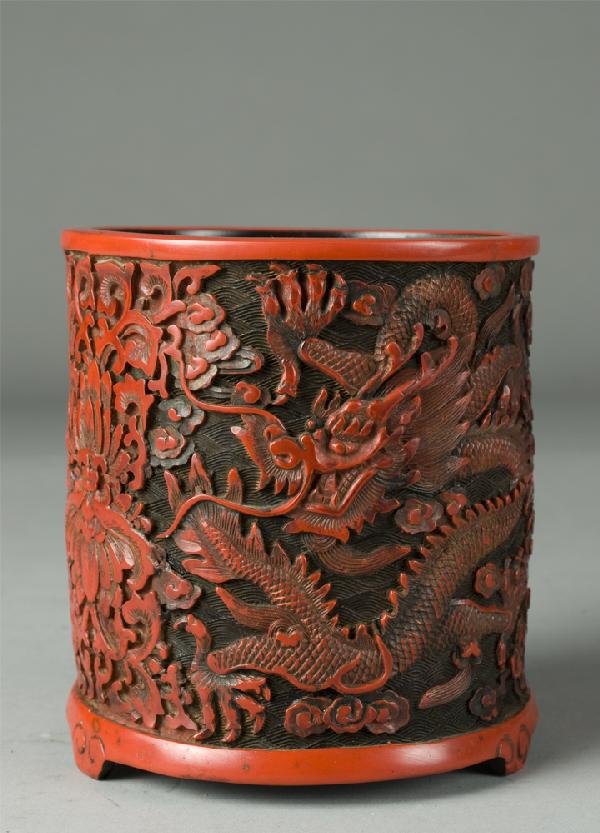 Finely Carved Cinnabar Brush Pot: Finely carved depicting two 5-clawed dragons amid ruyi clouds and separated by two lotus flower and scrolled swag designs, resting on three feet. 5.25" H x 4 & 7/8" Dia. 19th/20th Century.